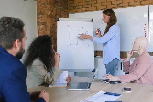 Group of pensive coworkers sitting at wooden table with gadgets and documents and listening presentations of colleague drawing graph in flipchart in modern office workspace in daytime. Social media training team roles: seven unranked functions